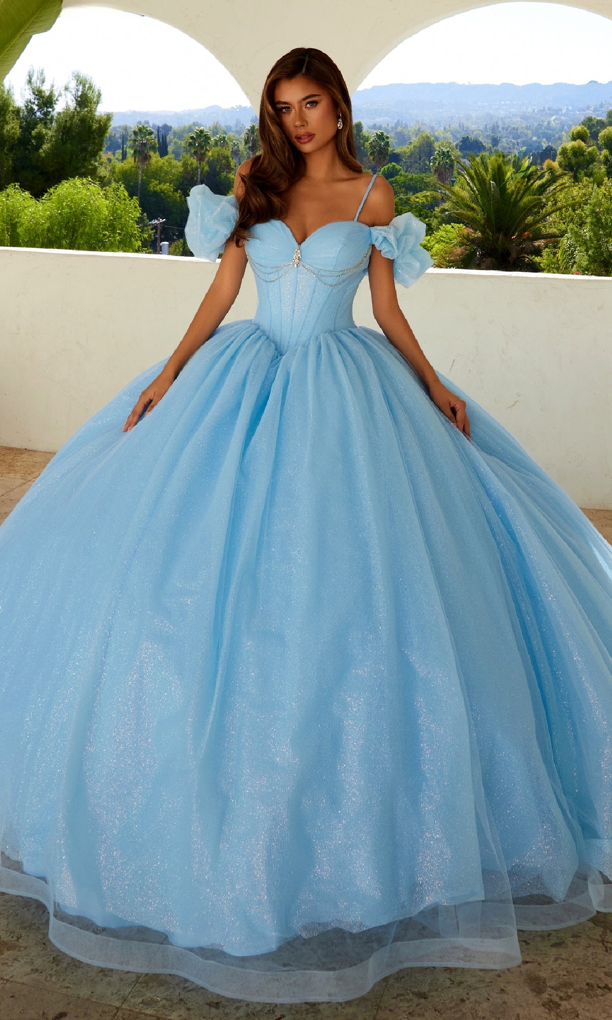 A woman stands on a patio in the Quinceanera Dress JT1487H By Juliet, a glitter tulle ball gown with off-the-shoulder puff sleeves and a fitted bodice. Green trees and hills in the background complete her princess look.